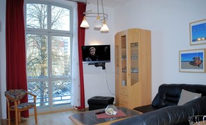 Una sala de estar con televisor, un mueble de madera, un sofá, un sillón y una ventana con cortinas rojas.