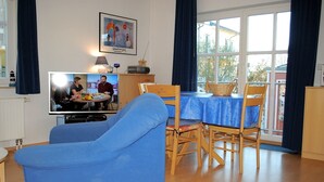 Ein Wohnzimmer mit Fernseher, blauem Sofa, Holzstühlen, einem Esstisch mit blauer Tischdecke und einem Fenster mit Vorhängen.
