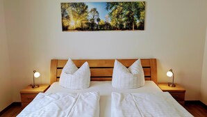 Een tweepersoonsbed met witte lakens, twee nachtkastjes met lampen, en een schilderij van een boslandschap erboven.