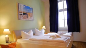 A double bed with white linens, a nightstand with a lamp, a painting of a beach scene, and a window with dark curtains. - Villa Anna an der Ahlbecker Strandpromenade, App. 05 (Ostseebad Heringsdorf OT Ahlbeck)