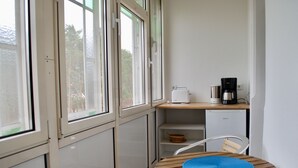Een kleine keuken met een witte koelkast, een koffiezetapparaat en een broodrooster op een houten aanrecht. Er staat een stoel met een blauwe mat en een wit kastje met een mandje.