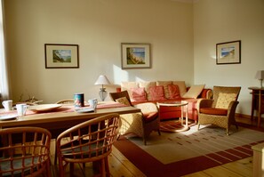 Ein gemütliches Wohnzimmer mit einem Holzesstisch, Korbstühlen, einer Couch mit orangen Kissen und gerahmten Landschaftsbildern an den Wänden.