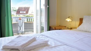 Ein Hotelzimmer mit einem Bett, einem Nachttisch mit Lampe, einem Sessel und Blick auf ein Gebäude durch eine Glastür.