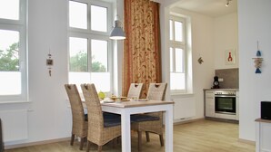 Spiseområde med et hvidt bord og fletstole, et køkken med moderne hårde hvidevarer, og store vinduer med gardiner.
