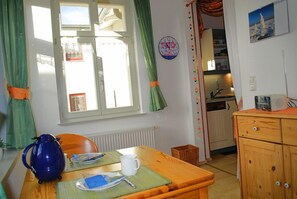 Une cuisine chaleureuse avec une table en bois, une bouilloire et une tasse. Une fenêtre avec des rideaux est visible, et une horloge est accrochée au mur.