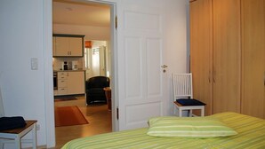 Een slaapkamer met een bed, een houten garderobe, een stoel en uitzicht op de keuken.
