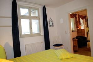 Une chambre à coucher comprenant un lit, une fenêtre, un radiateur, une chaise et une porte donnant sur une autre pièce.