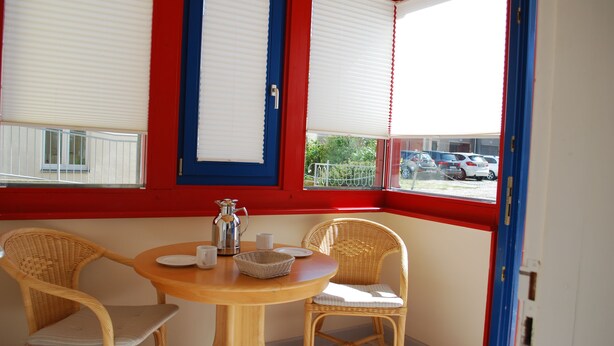 Una habitación con una mesa redonda de madera, dos sillas de mimbre y un ventanal con adornos rojos y azules.