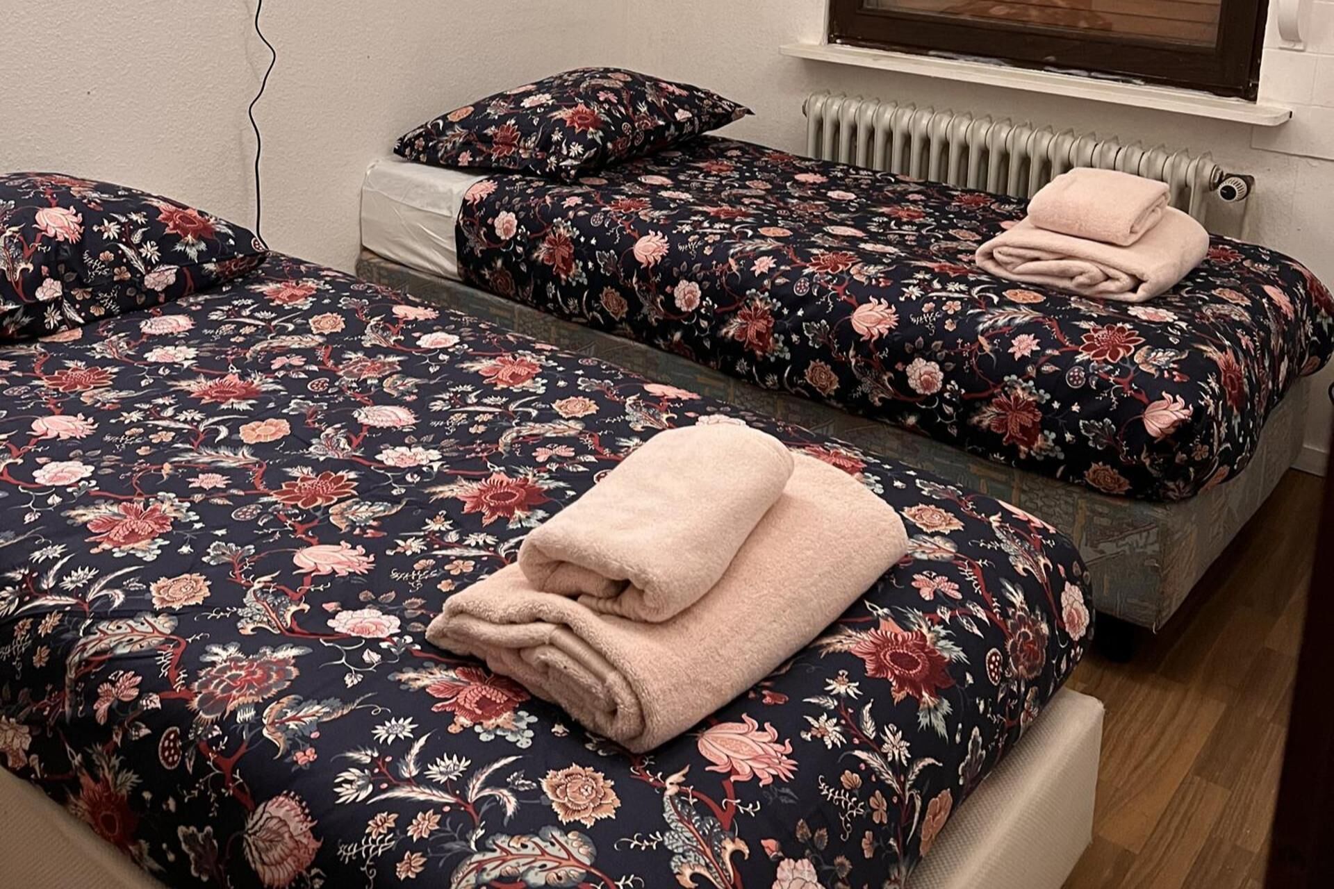 A room with two beds, each with a floral-patterned bedspread and matching pillows, and a radiator above one bed.