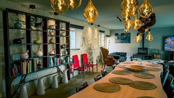 Een ruime woonkamer met een lange houten tafel, rode stoelen, een boekenplank vol diverse spullen en moderne hanglampen.