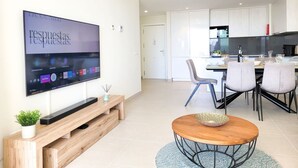Een moderne woonkamer met een flatscreen televisie aan de muur, een houten tv-meubel, een ronde salontafel en een eethoek, met een keuken op de achtergrond.