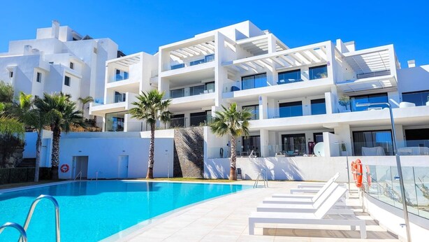Een modern hotel met een zwembad, meerdere balkons en een helderblauwe lucht.