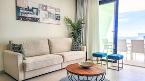 Een moderne woonkamer met een beige bank, een ronde salontafel en een groot raam met uitzicht op zee.
