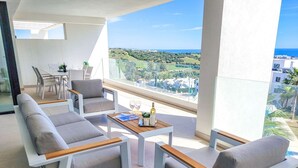 Een balkon met een zithoek, een eettafel en uitzicht op een kustlandschap.
