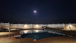 Un'area piscina di notte, con recinzione bianca, sedie a sdraio e una luna nel cielo.