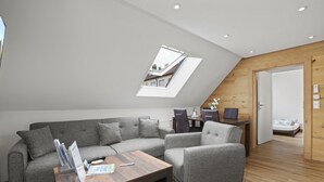 Una sala de estar moderna con un sofá seccional gris, una mesa de centro de madera y una mesita con revistas y un control remoto. Hay un tragaluz sobre el sofá y una puerta de madera al fondo.