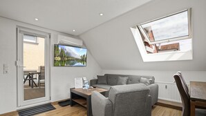 Un salón moderno con un sofá gris, una mesa de centro y una mesa de comedor. Hay un ventanal con vistas a una montaña y un lago.