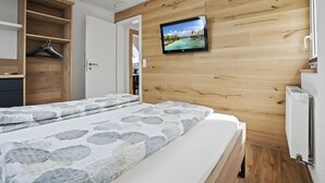 Un dormitorio con una cama, un armario de madera, un televisor montado en la pared y una puerta que da a otra habitación.