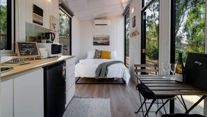 Une chambre compacte et moderne, dotée d’une kitchenette, d’un lit avec une literie blanche, d’un coin repas avec une table et des chaises, et de grandes fenêtres offrant une vue sur l’extérieur.