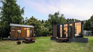 Dos cabañas modernas y compactas con exteriores de madera y ventanas amplias, ubicadas en un área verde con árboles al fondo.