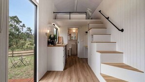 Cocina compacta con piso de madera, armarios blancos y un pequeño comedor visible a través de una puerta abierta.