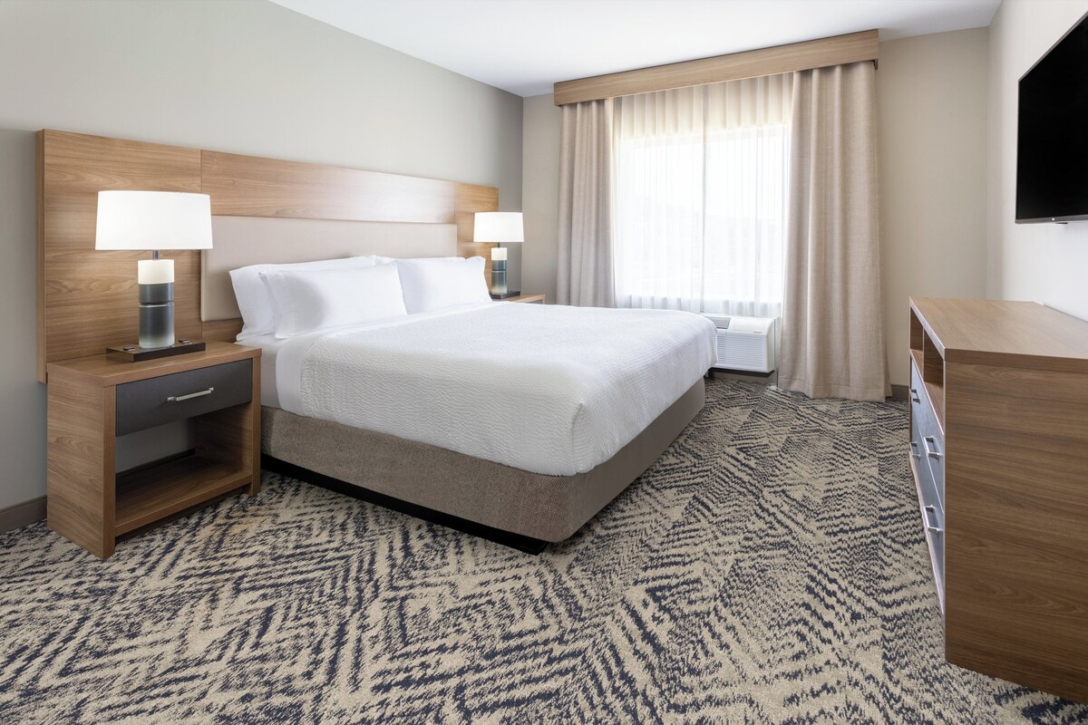 Une chambre d’hôtel comprenant un lit, deux tables de chevet avec lampes, un téléviseur à écran plat, une fenêtre avec rideaux et un sol recouvert de moquette.