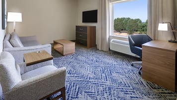 A hotel room with a sofa, a desk, a chair, a TV, and a window with a view of trees.