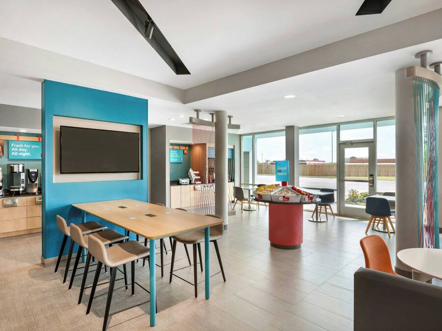 A modern hotel lobby with a large flat-screen TV, a coffee station, and a seating area with tables and chairs.