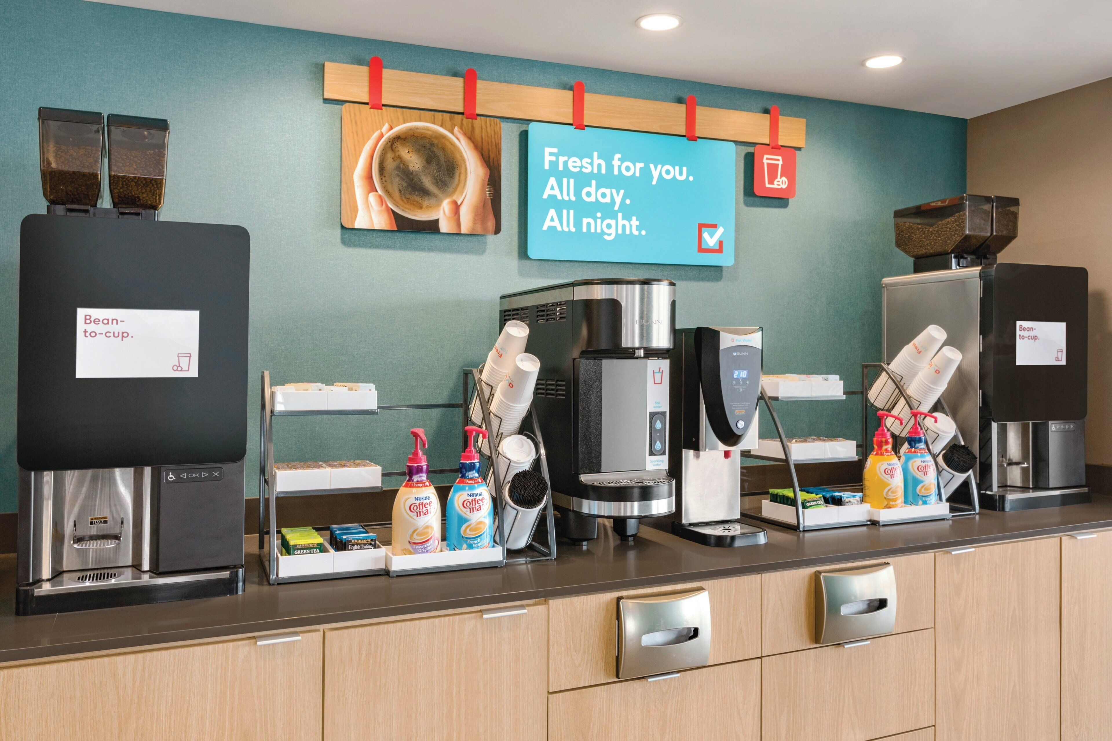 A coffee station with multiple coffee machines, a sign indicating fresh coffee availability, and dispensers for coffee creamer and milk.