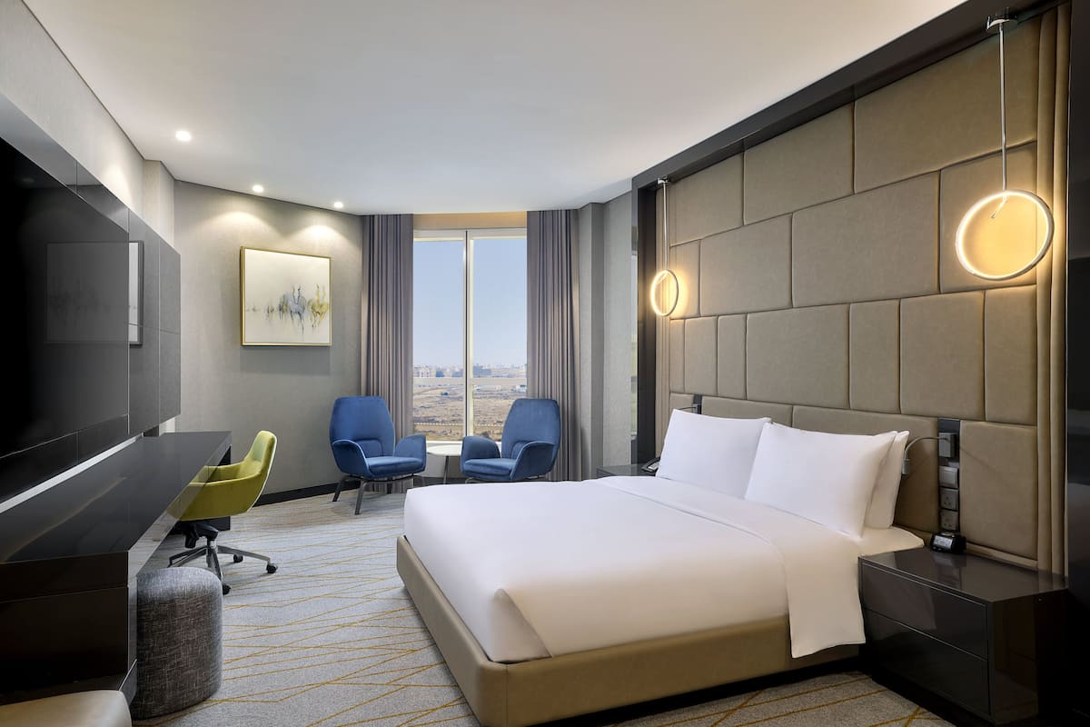 Ein Hotelzimmer mit einem großen Bett, einem Schreibtisch, zwei Sesseln, einem Fernseher und Blick auf die Stadt.