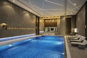 Une piscine intérieure moderne dotée d’un plafond au design géométrique, de chaises longues et offrant une vue sur l’espace réservé au restaurant.
