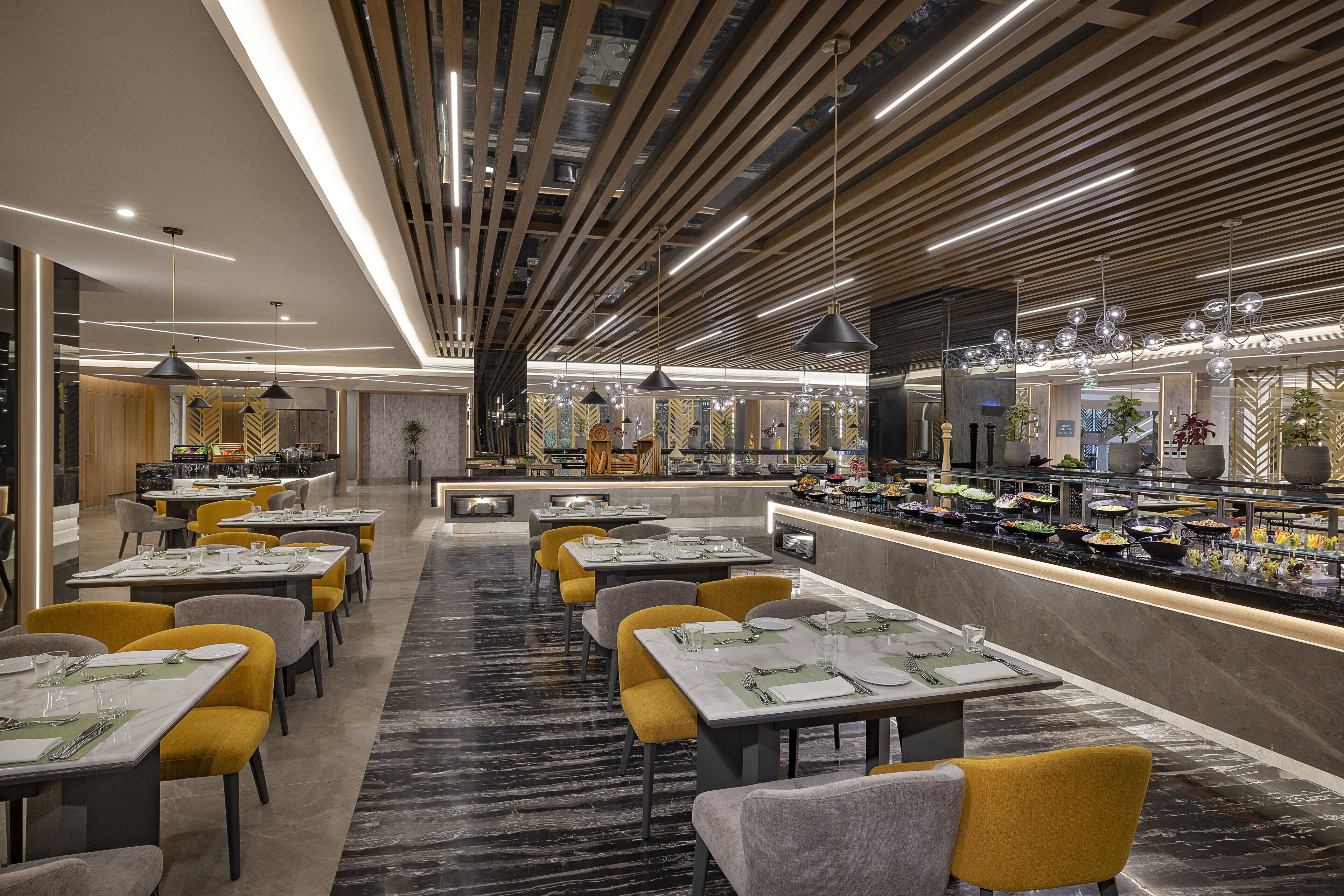 Restaurante moderno com mesas compridas, cadeiras amarelas e uma variedade de pratos expostos no balcão.