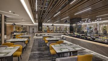 Restaurante moderno com mesas compridas, cadeiras amarelas e uma variedade de pratos expostos no balcão.