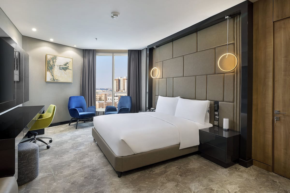 Ein modernes Hotelzimmer mit einem großen Bett, einem Schreibtisch, zwei Sesseln, einem Fernseher und Blick auf die Stadt.