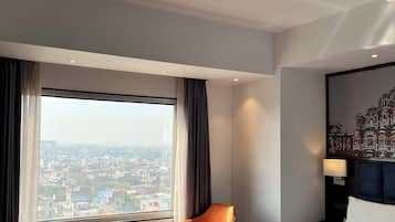 A hotel room with a large bed, a chair, a small table, and a view of the city through a large window.