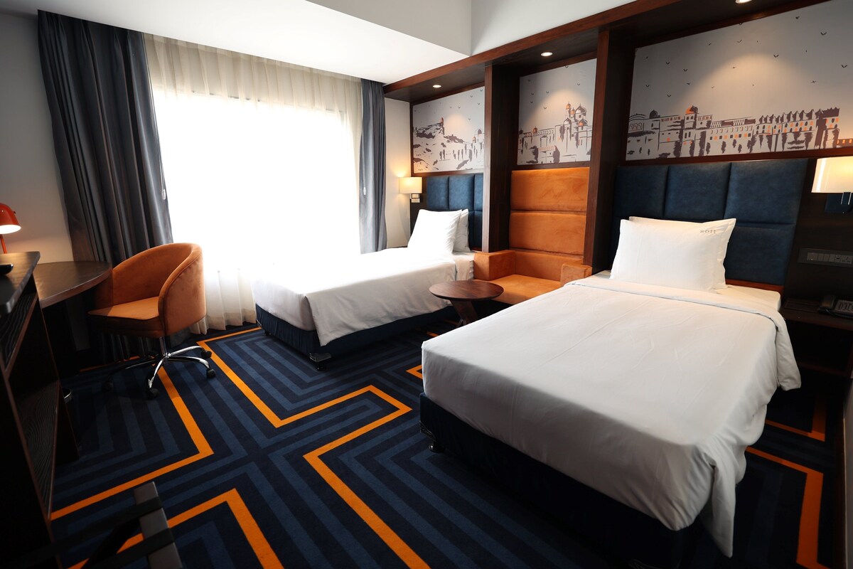 A hotel room with two beds, a desk, a chair, and a large window with curtains.