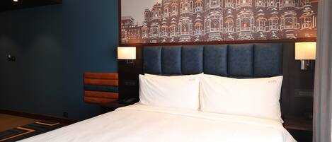 Quarto de hotel com uma cama grande, cabeceira, duas luminárias de mesa, uma obra de arte na parede e uma mesinha de cabeceira com um telefone.