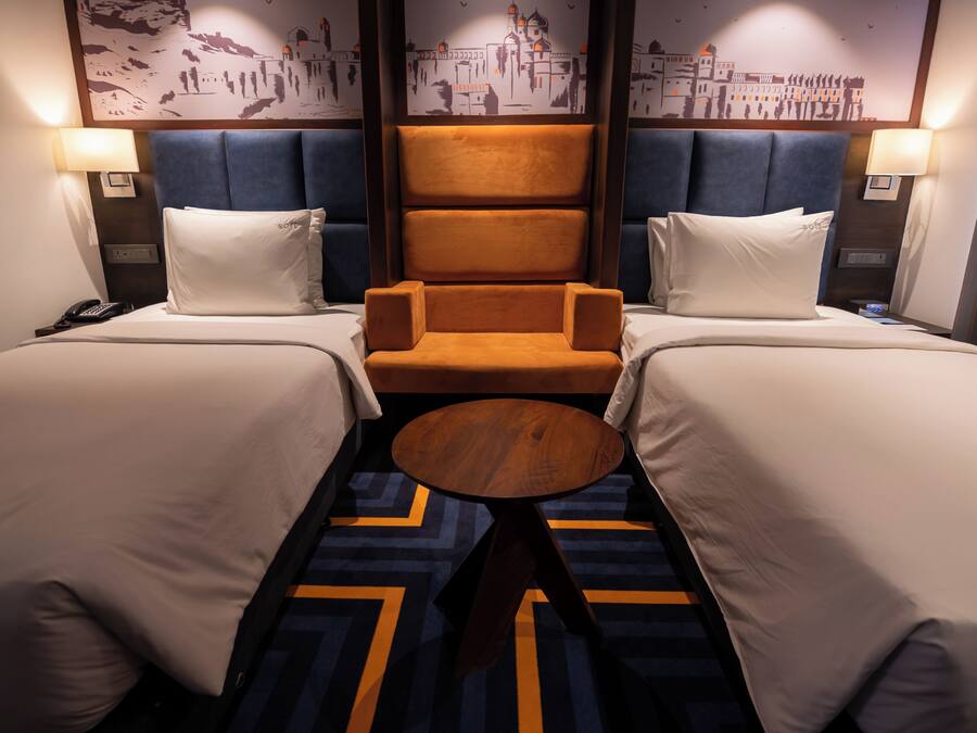 A hotel room with two beds, a central armchair, a small round table, and a cityscape mural on the wall.
