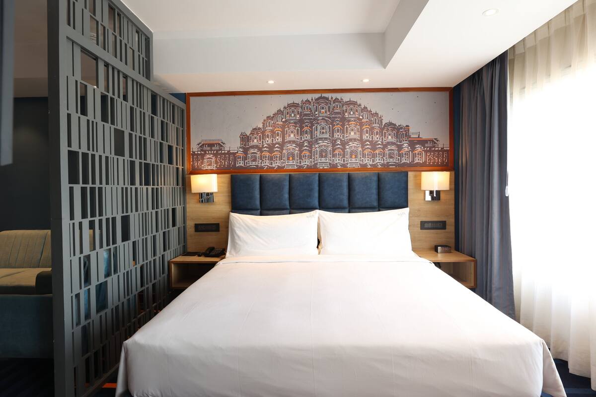Une chambre d’hôtel avec un grand lit, une fresque murale décorative, une tête de lit, deux tables de chevet avec des lampes et une fenêtre avec des rideaux.