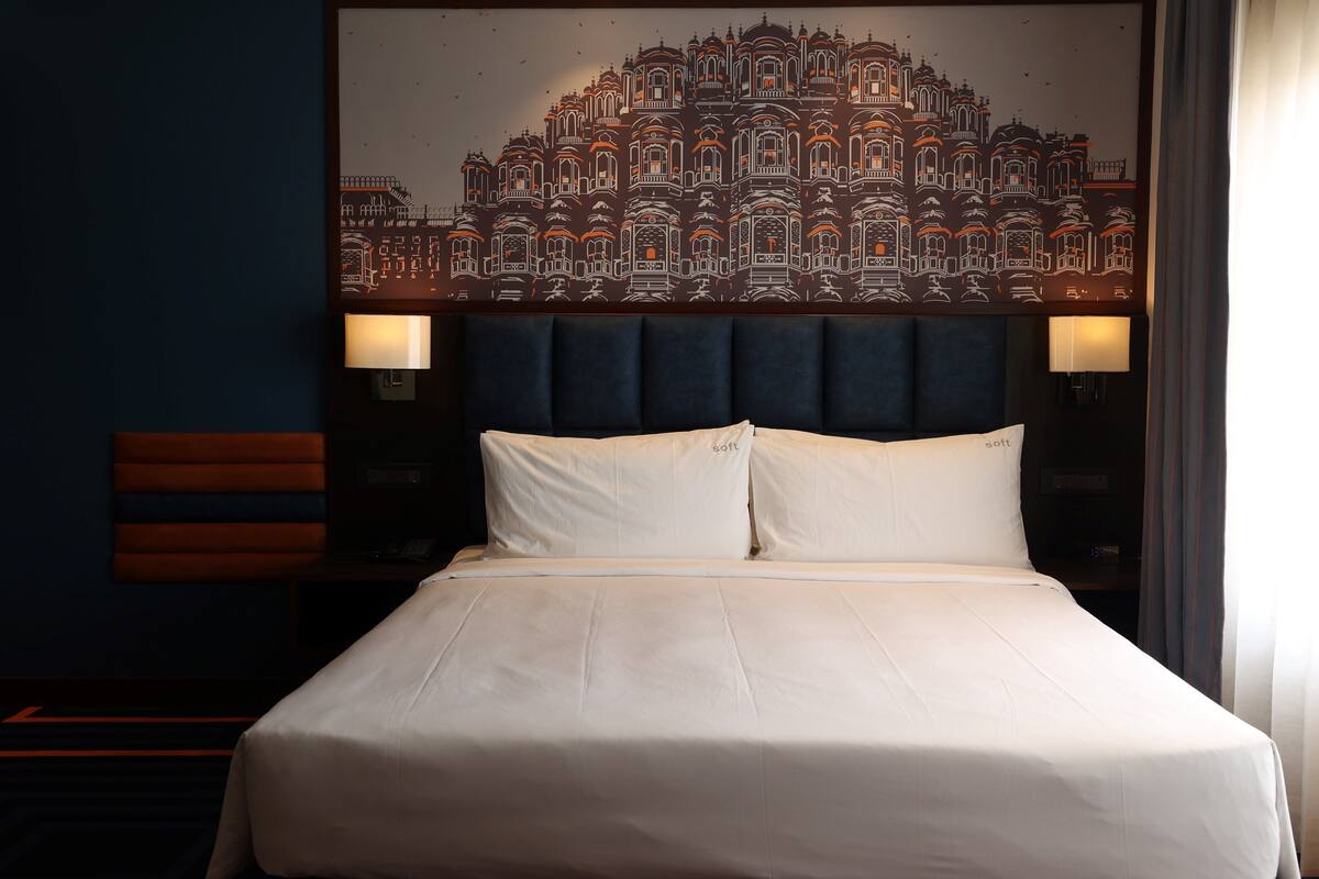 Une chambre d’hôtel avec un grand lit, deux lampes de chevet, une tête de lit ornée d’une fresque représentant un paysage urbain et une table de chevet avec une lampe.