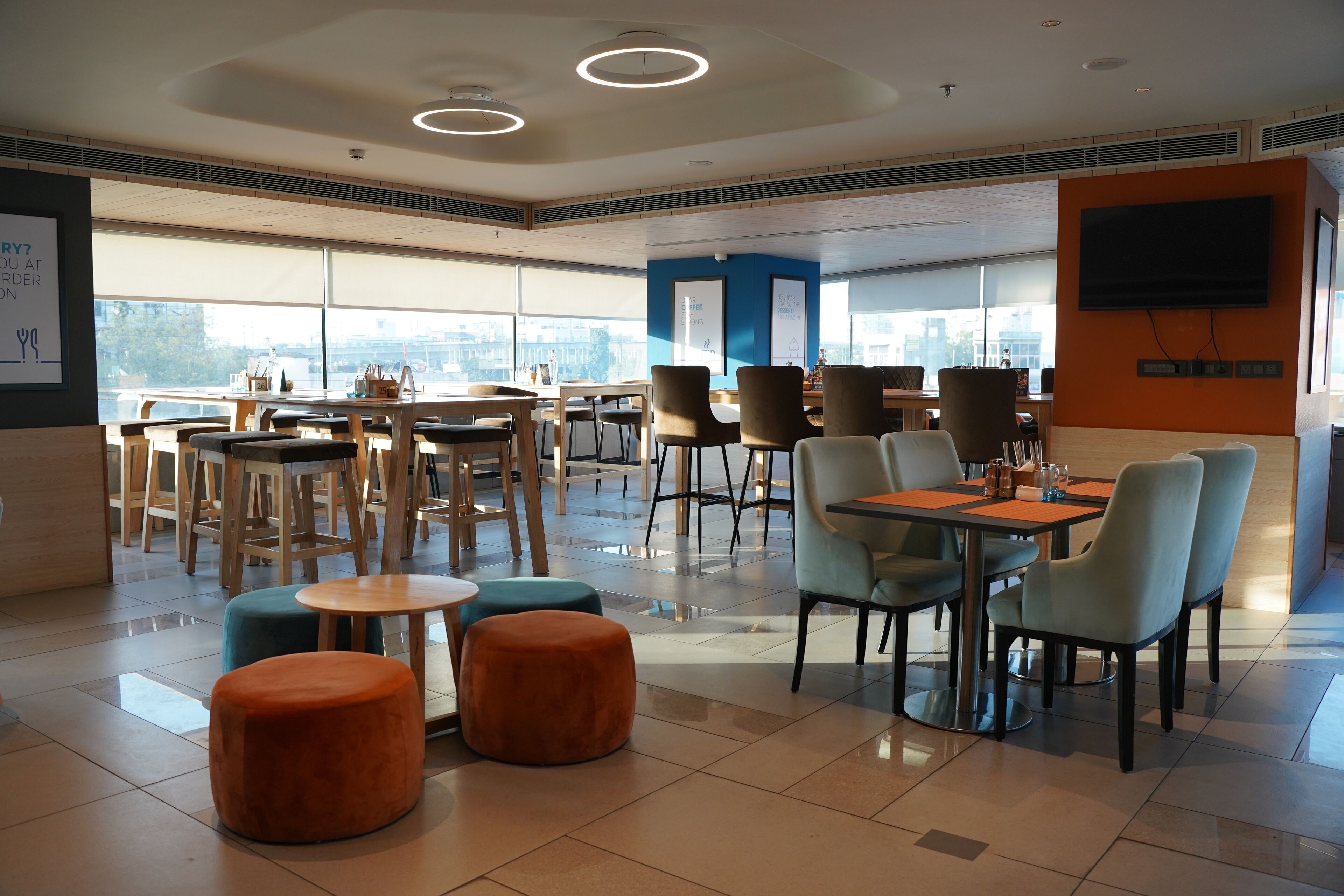 A modern dining area with multiple tables and chairs, large windows, and a flat-screen TV mounted on the wall.