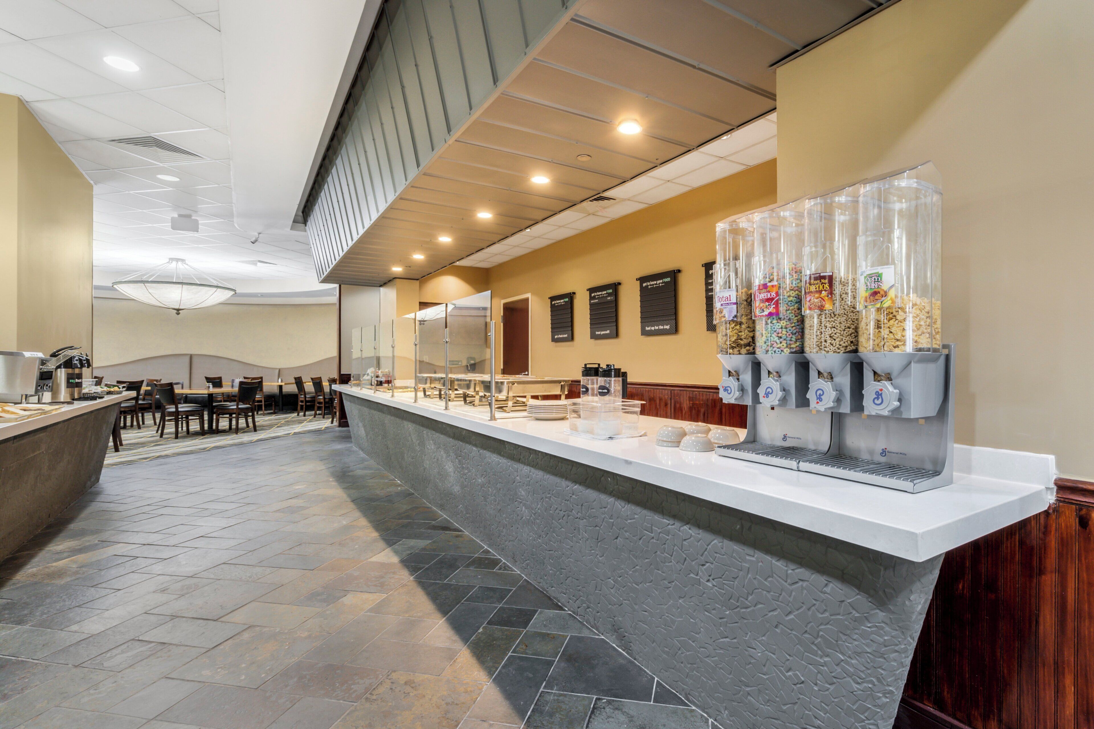 A hotel breakfast area with a long counter, multiple beverage dispensers, and seating arrangements.