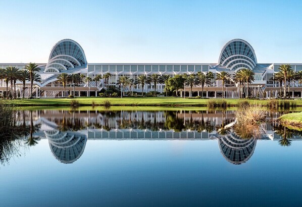 A modern hotel with a large reflective pool in front, palm trees, and a clear blue sky. - Holiday Inn Orlando International Dr-ICON by IHG (Orlando)
