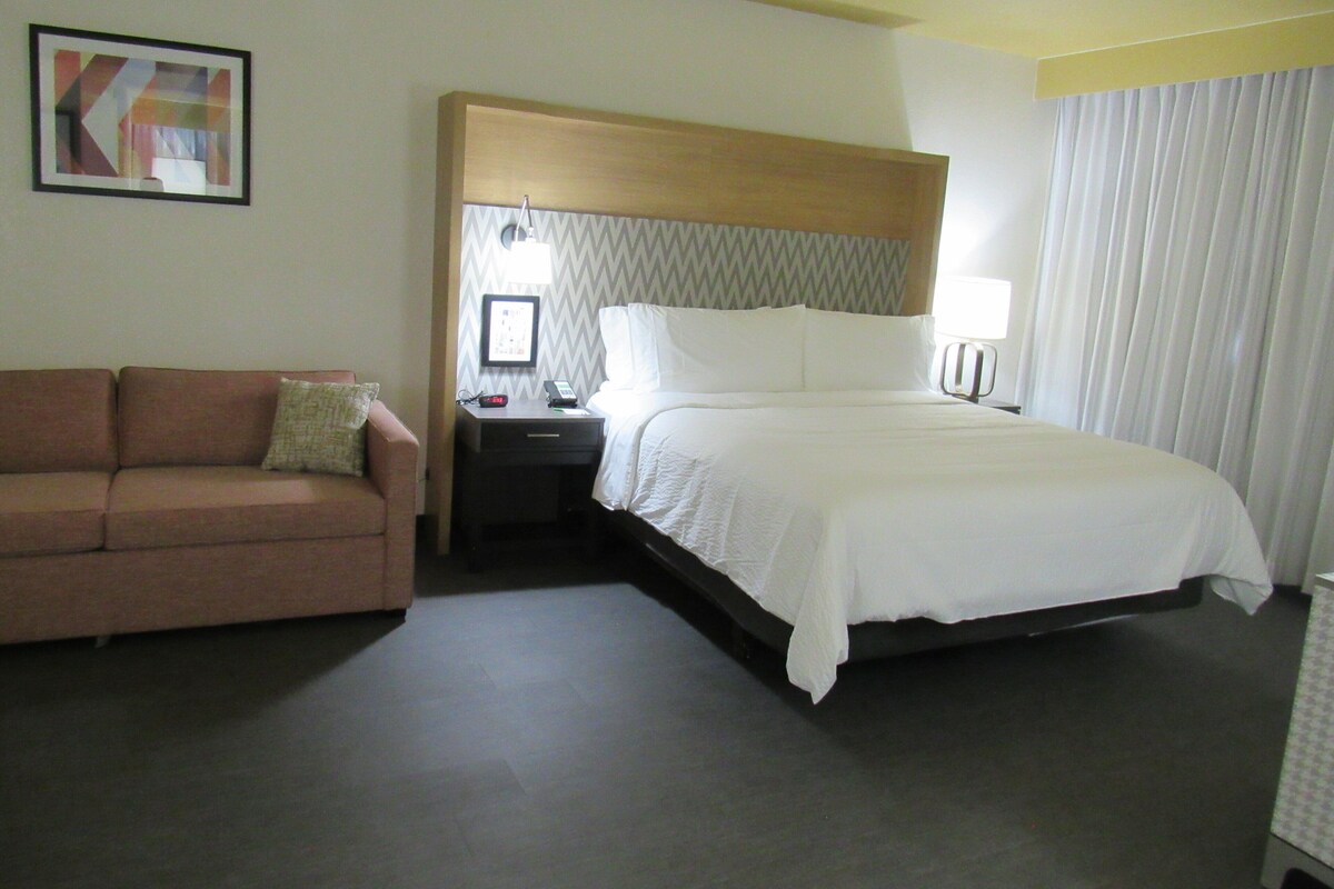 A hotel room with a bed, a nightstand, a lamp, a sofa, and a framed picture on the wall.