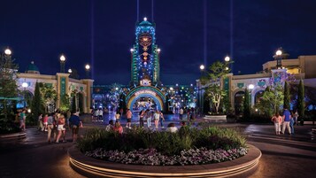 A nighttime view of a large, illuminated building with a clock tower, surrounded by a well-lit plaza with people and a flower bed.