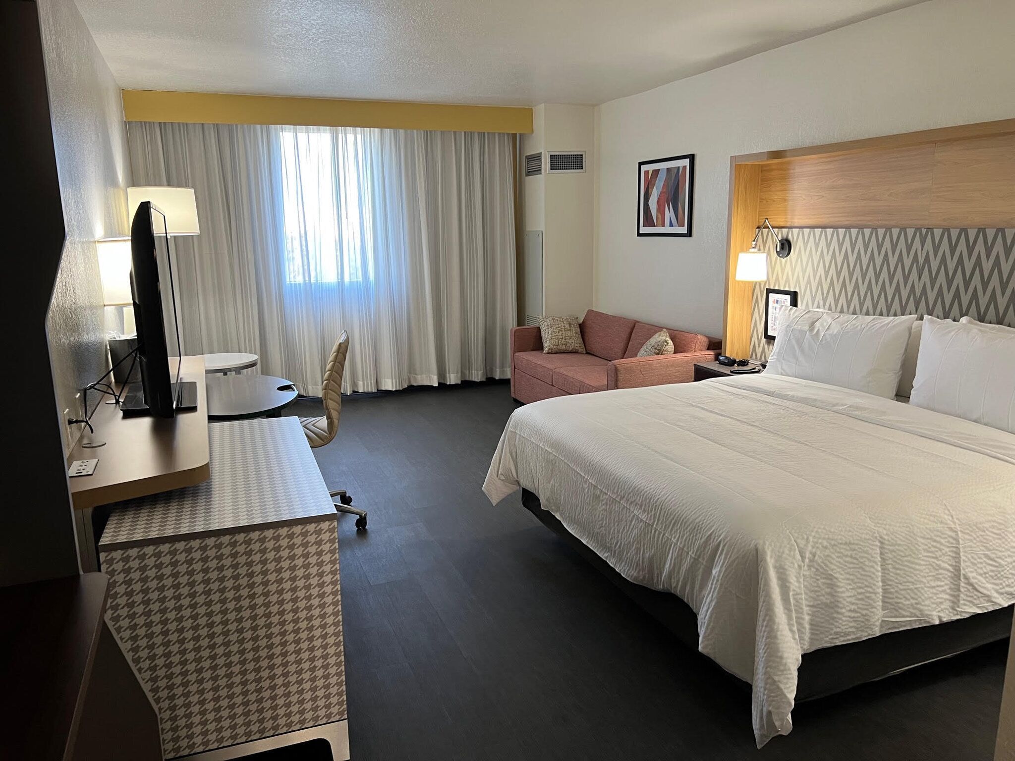 A hotel room with a bed, a desk with a computer, a chair, a sofa, and a window with curtains.