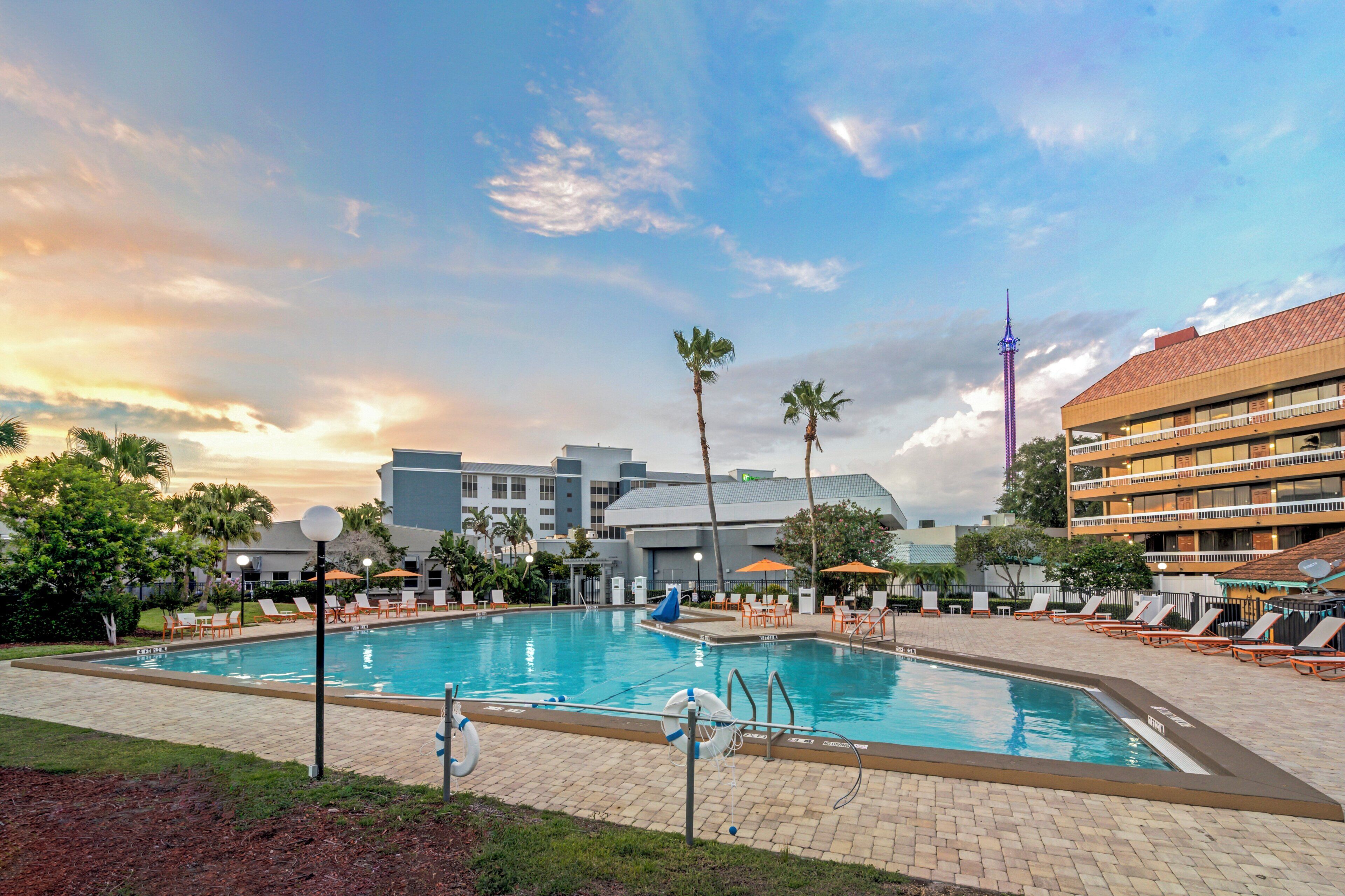 A swimming pool with lounge chairs and umbrellas, surrounded by palm trees and buildings.