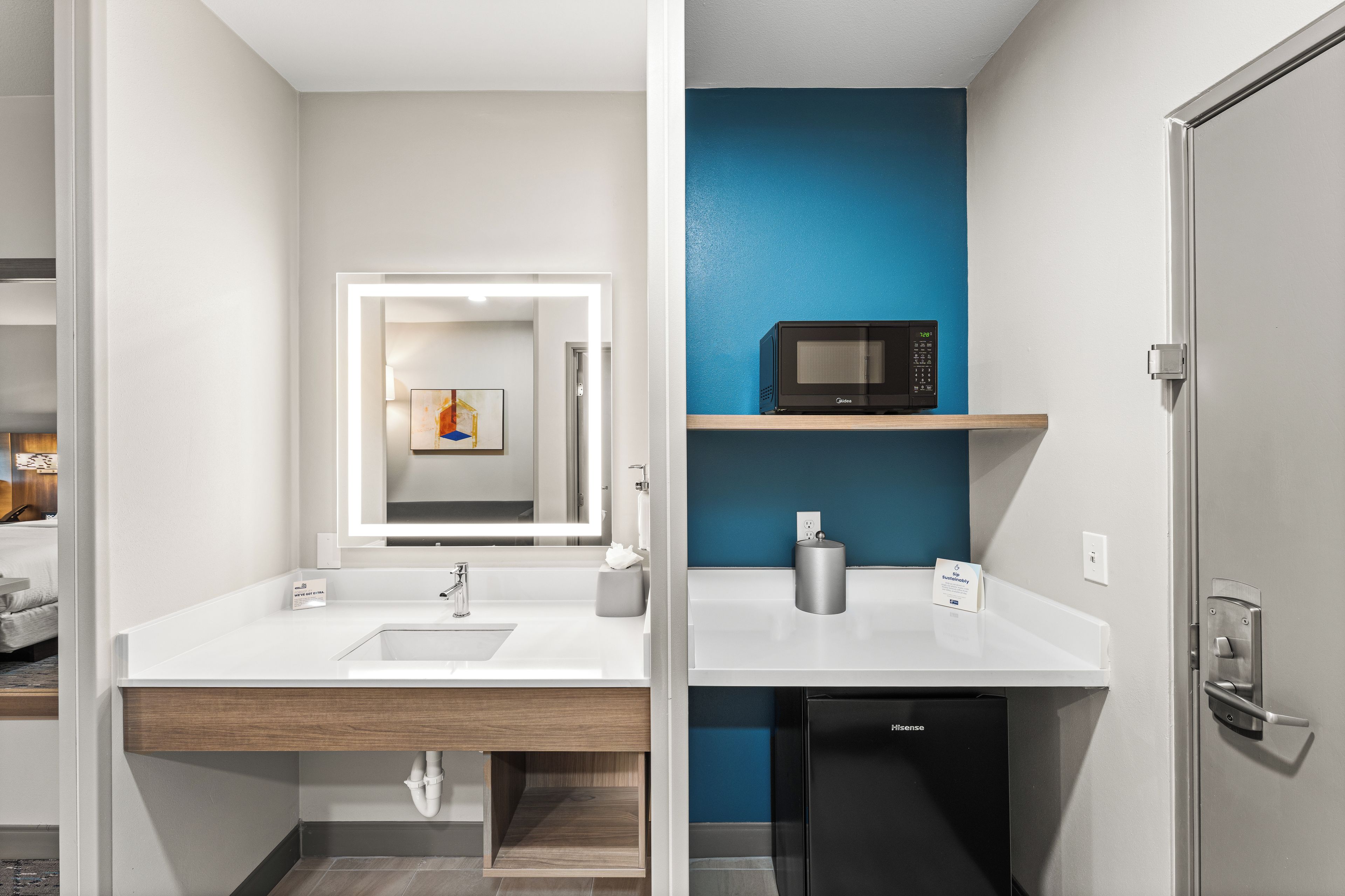 Un baño moderno con un lavamanos blanco, un espejo, un microondas y un refrigerador negro.