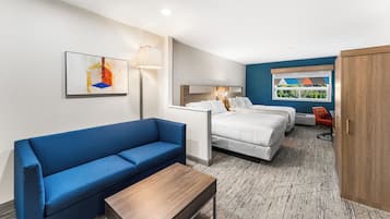 Habitación de hotel con sofá azul, una mesa de madera, dos camas, una mesita de noche, una lámpara y una ventana con vistas.