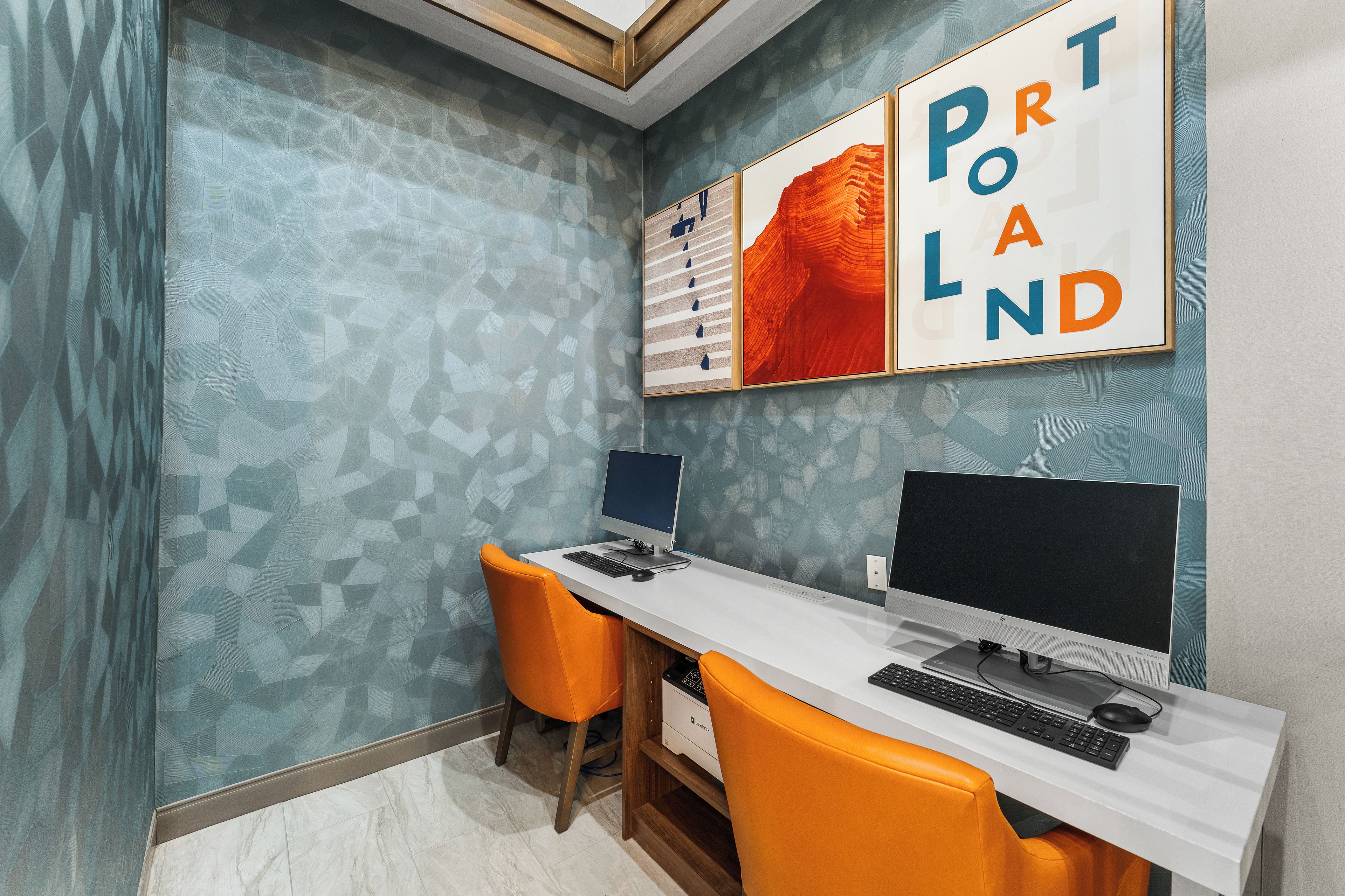 A modern office space with two desks, each with a computer and keyboard, and two orange chairs. The walls have a geometric pattern, and there are framed artworks above the desks.